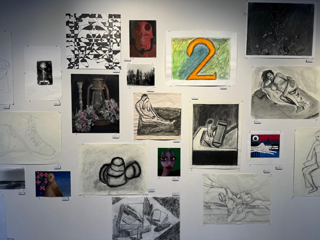 collection of student art on gallery wall. My favorite is a colored charcoal drawing of a lamp with a candlestick and roses around it. It feels bleak and drained of life. Although it is in color, the colors are muted for effect.